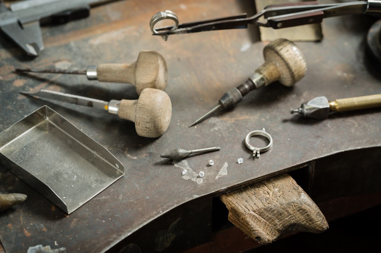 A close-up of a jeweler’s table with different tools and an engagement ring setting with several diamonds scattered nearby.