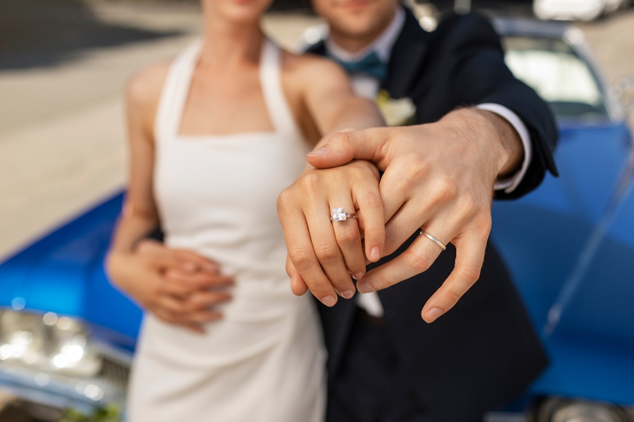 A close-up of a newlywed couple standing in front of the blue car, showcasing their wedding band and engagement ring.