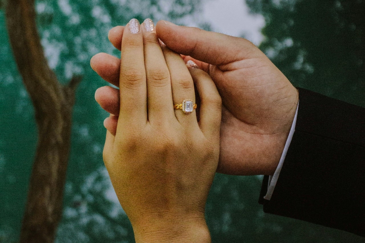 A close up of a two hands held together, showcasing yellow gold emerald cut diamond engagement ring with intertwined band against natural backdrop.