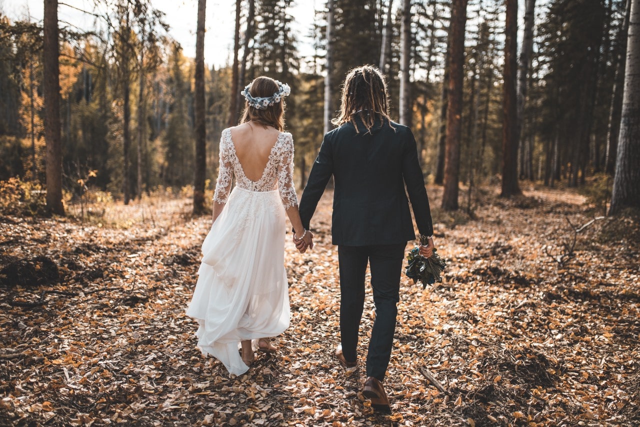 Bride with open back lace wedding dress and groom in the dark grey suit walk together hands by hands in the woods full of fallen leaves.