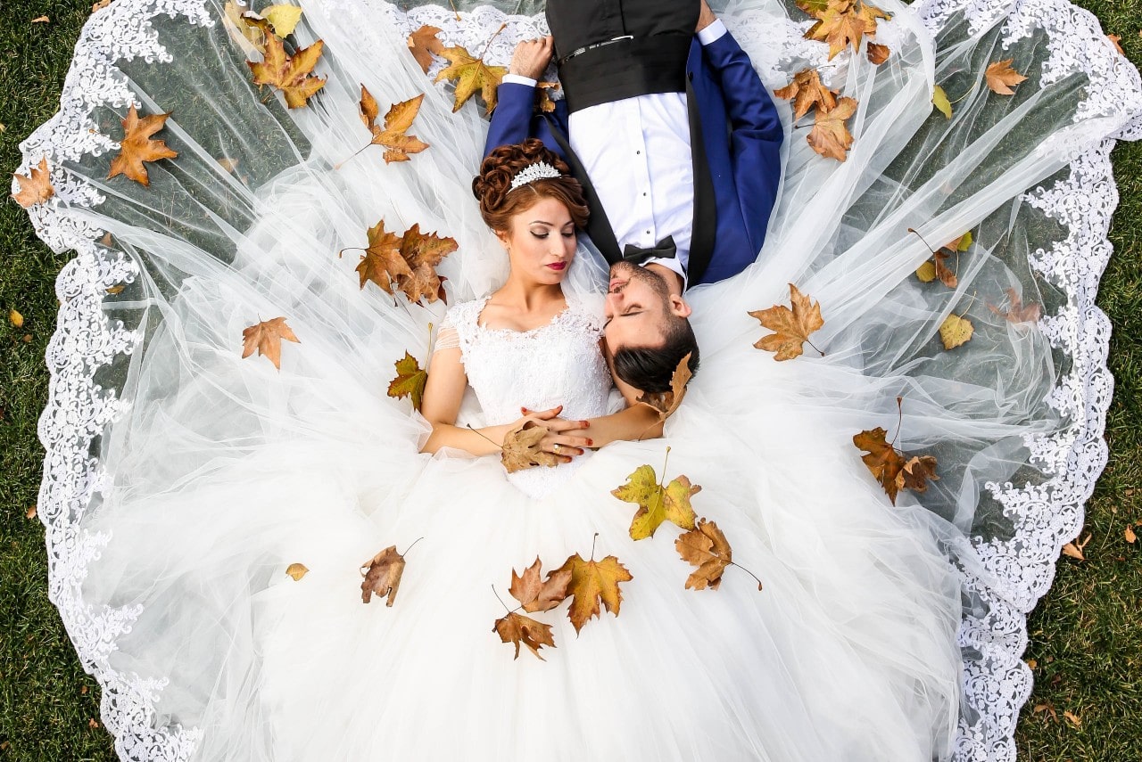 Bride and groom lie on grass, embraced, her white lace gown spread like a fan. Autumn leaves are scattered around them, creating a serene and romantic mood.