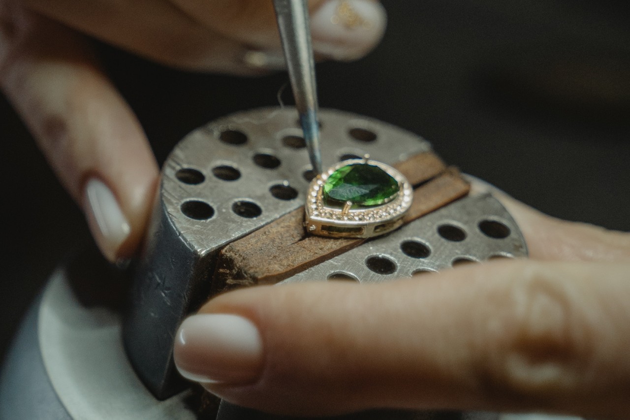 A close-up of a jeweler&rsquo;s hands as they work on an intricate emerald and diamond pendant.