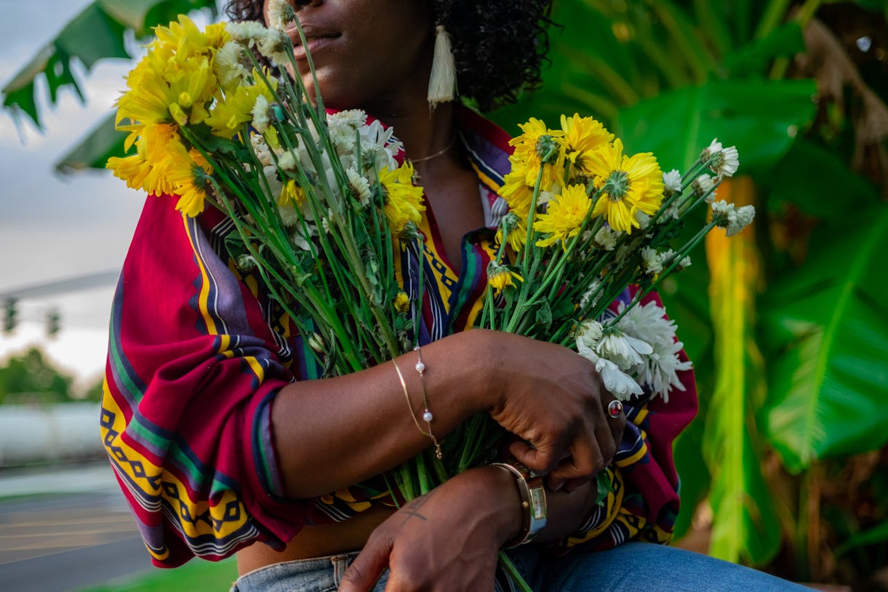 A close-up of a woman in a vibrant blouse holding a colorful bunch of flowers in front of palm leaves.
