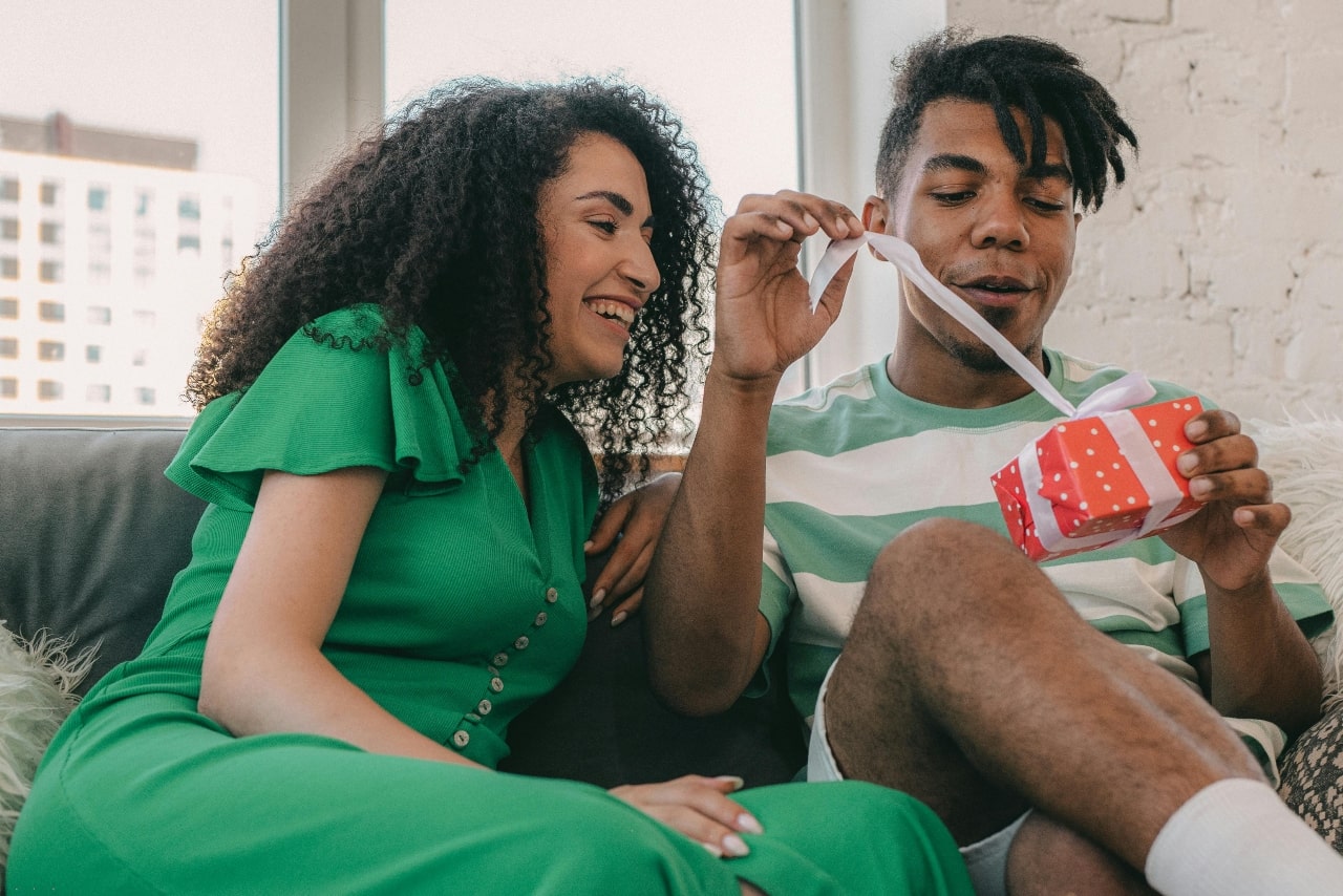 A young man opens a small red gift box with a ribbon while sitting beside a smiling woman in a green dress.