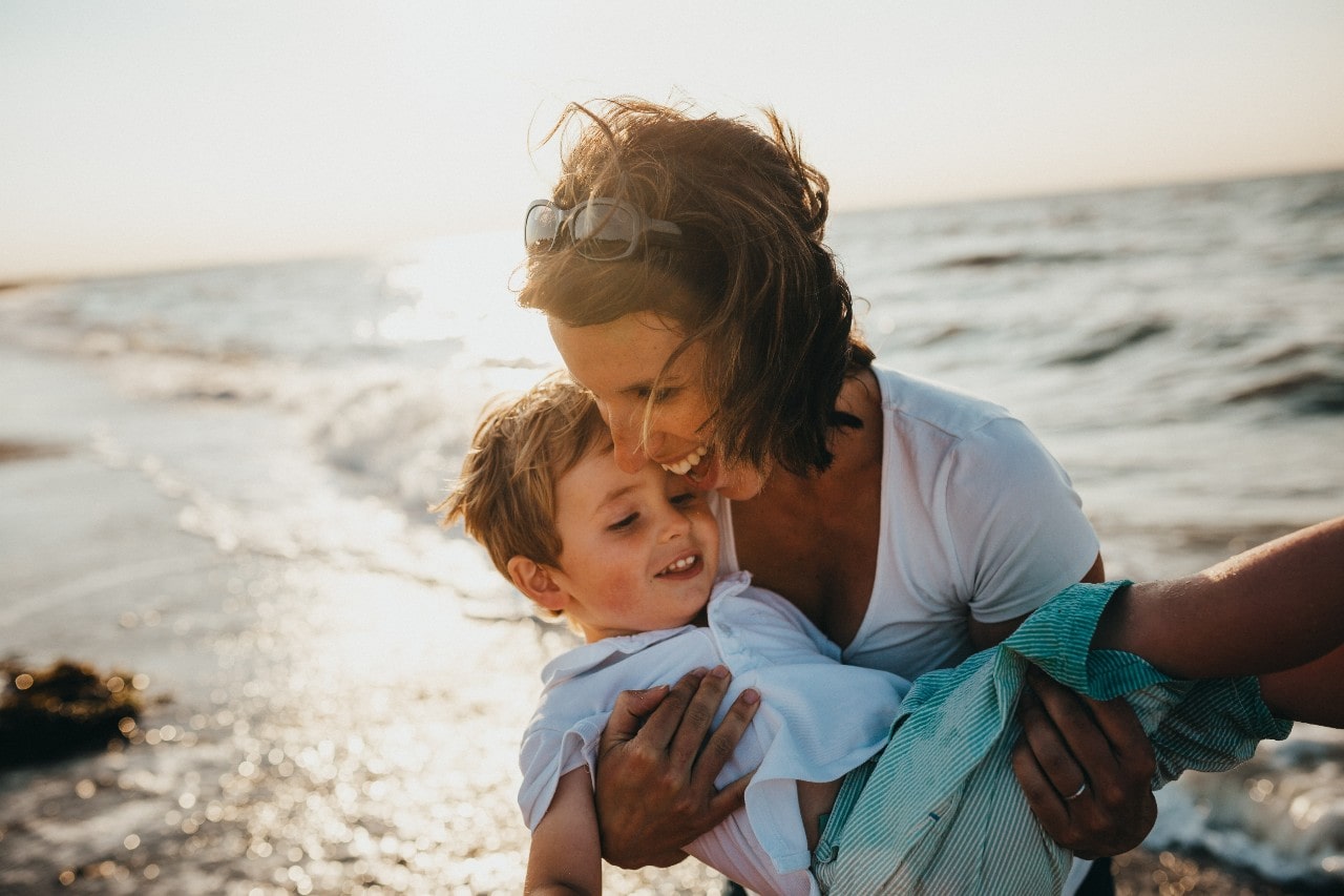 A picture of a mother with short hair holding her son on sunlight seashore.