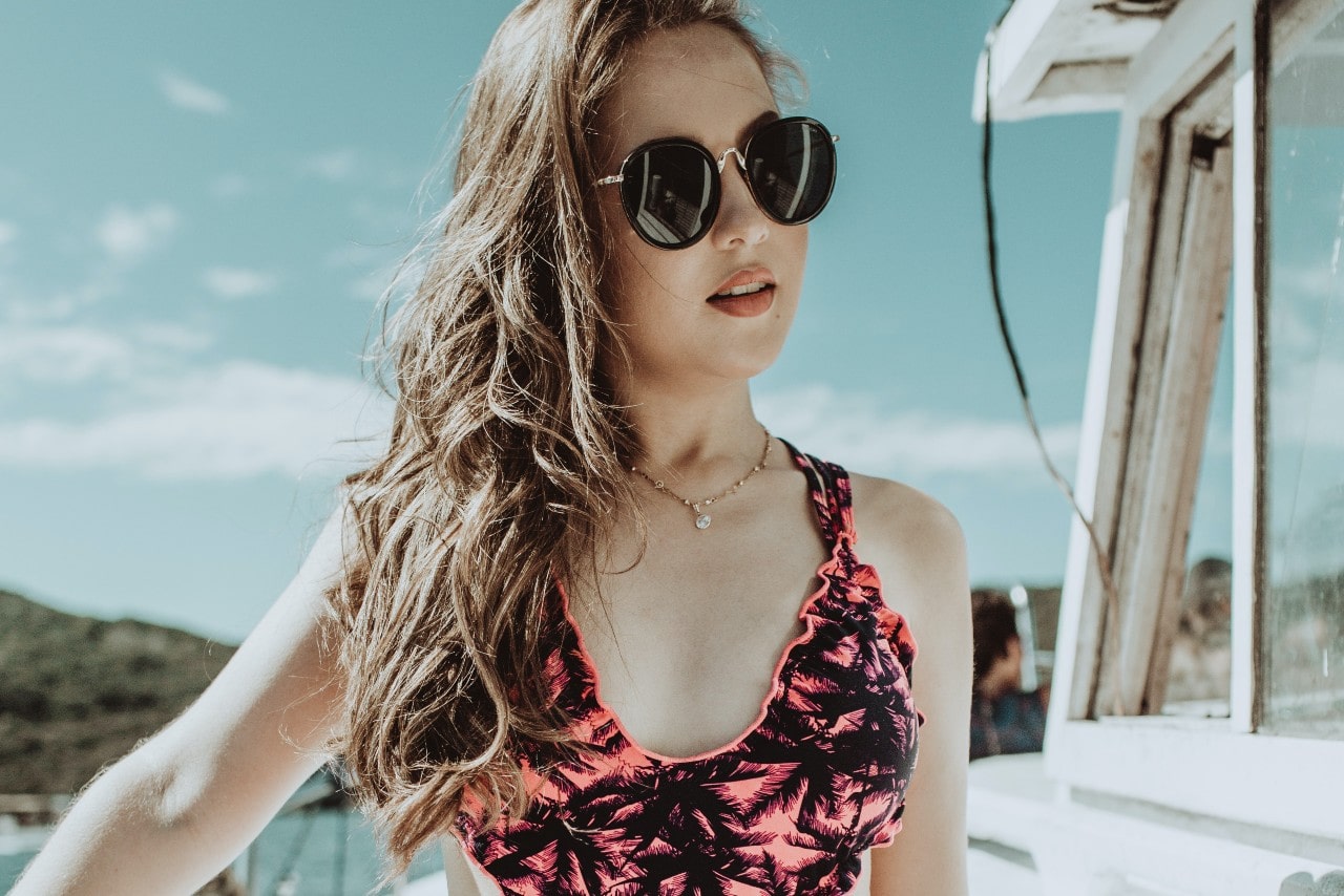 A young woman in sunglasses and a pink palm-patterned swimsuit poses confidently outdoors on a sunny day.