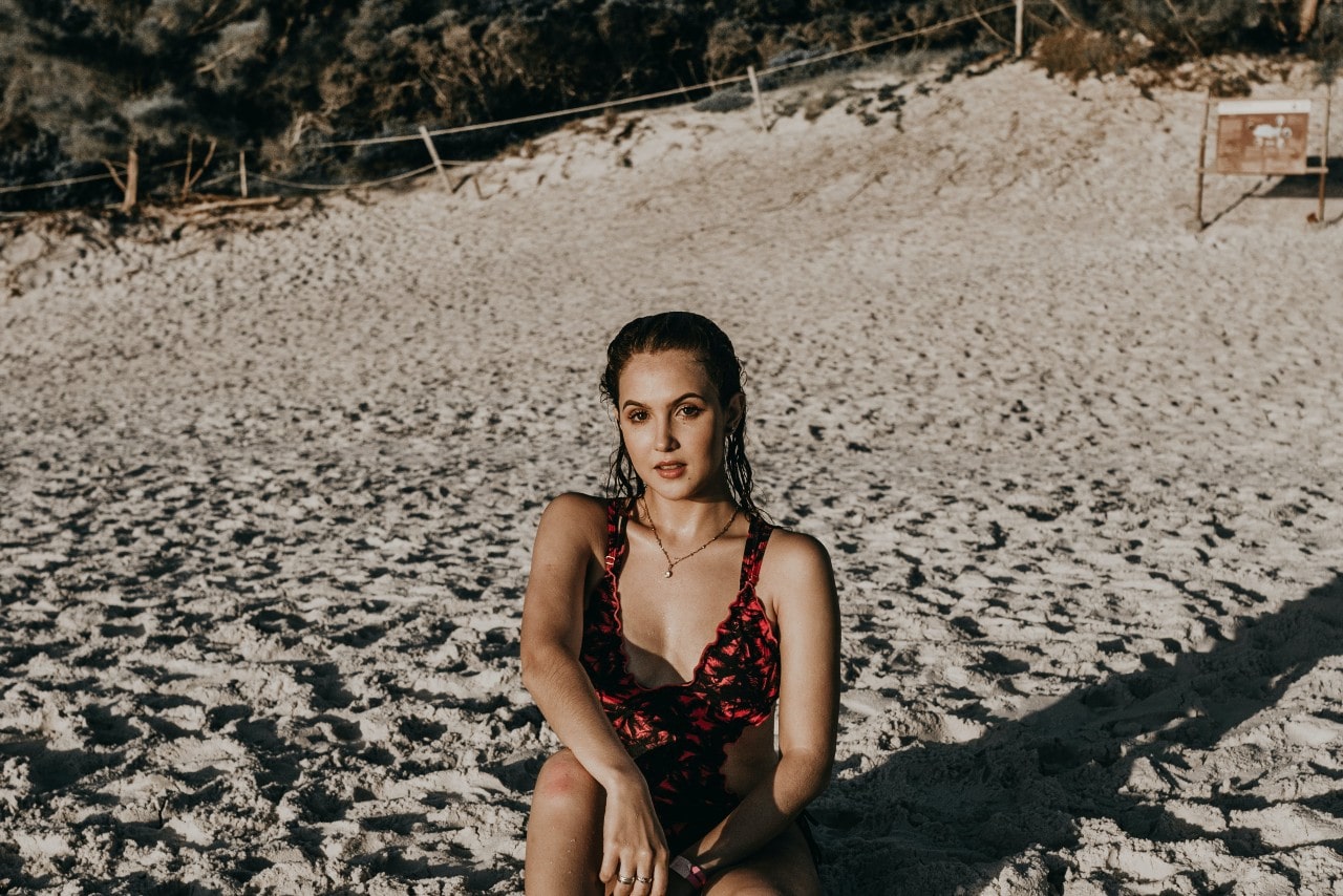 A woman in a red swimsuit sits on a sandy beach wearing a pendant.