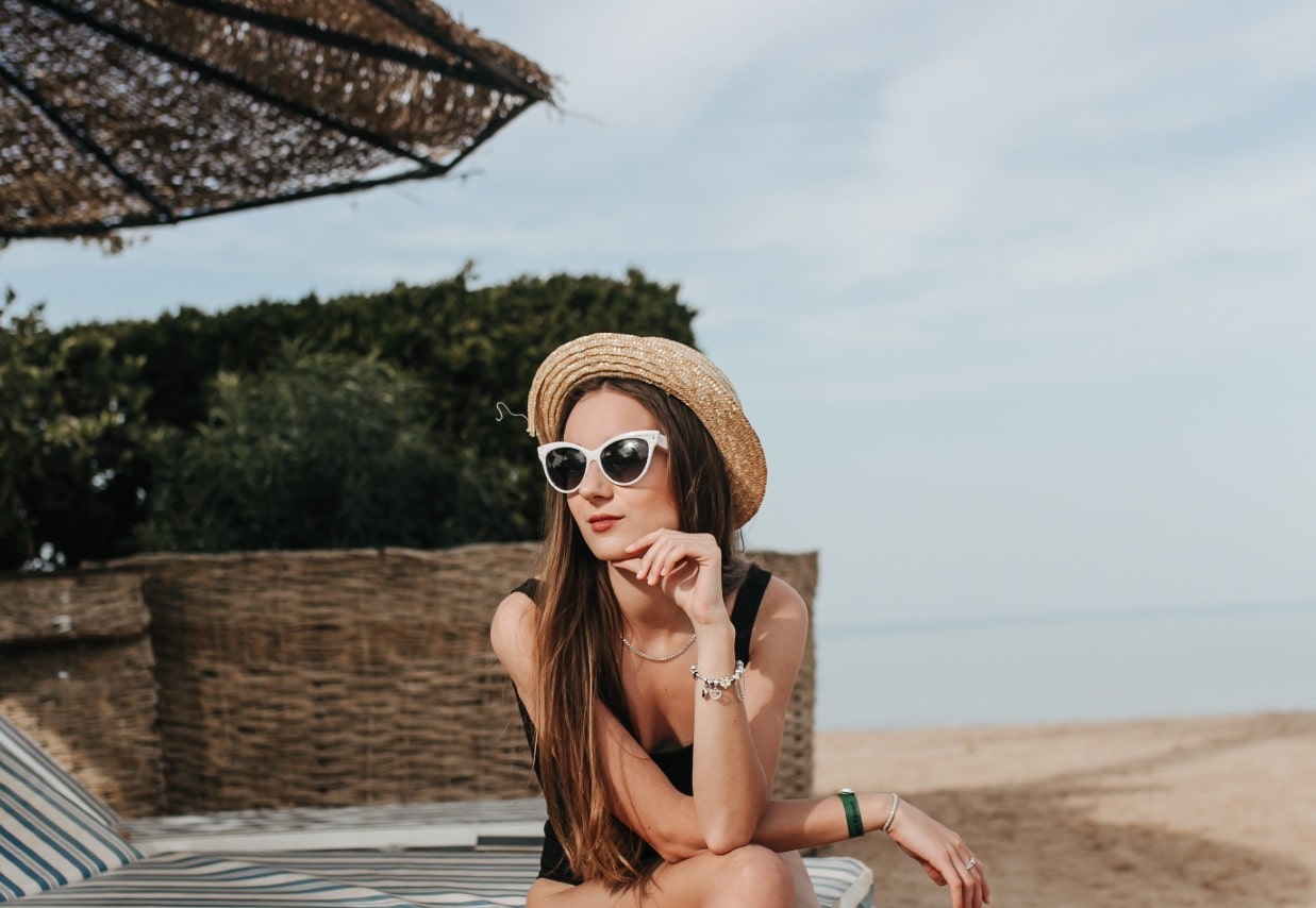 A woman in a straw hat and sunglasses and wearing fashion jewelry sits on a beach chair.