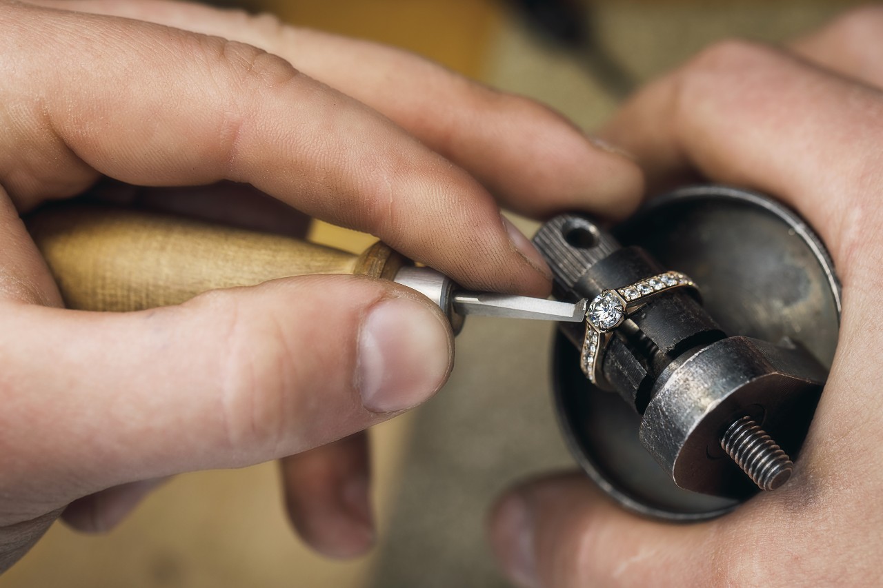 A close-up of hands adjusting a diamond ring with a tool on a jeweler's vice.