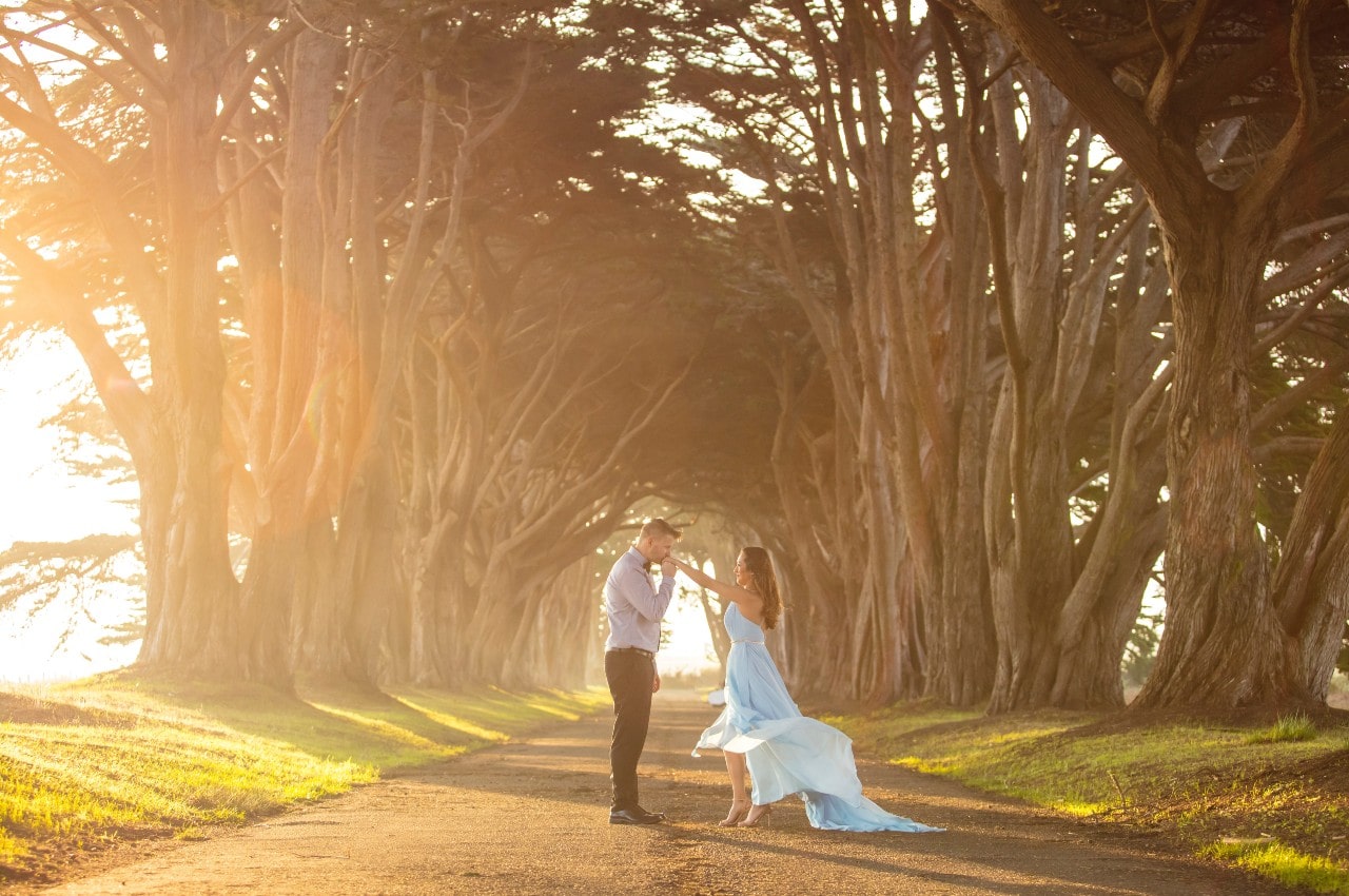 A man kissing a woman&rsquo;s hands as her dress blows in the breeze, standing on a trail surrounded by trees and sunshine.