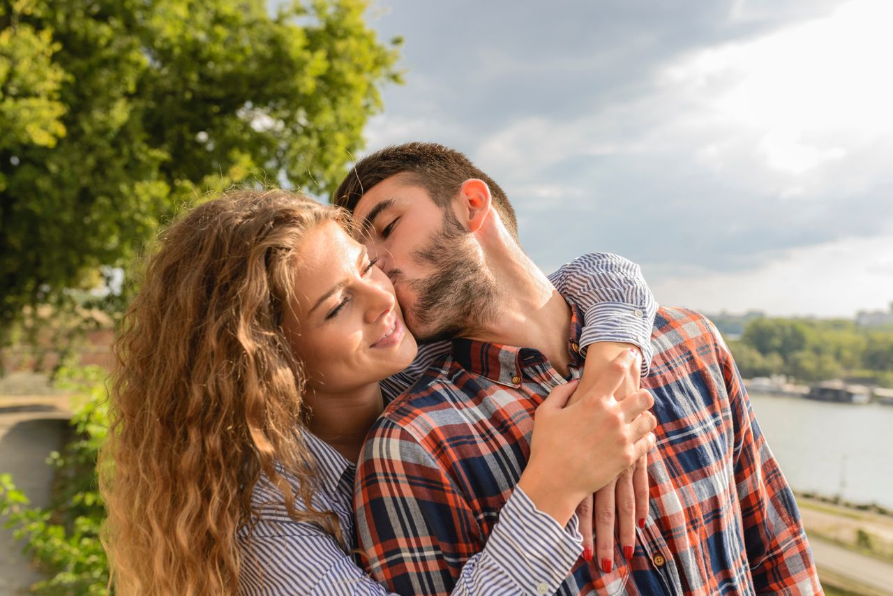 A woman wrapping her arms around her partner, he leans in and kisses her cheek, surrounded by a nature scene.
