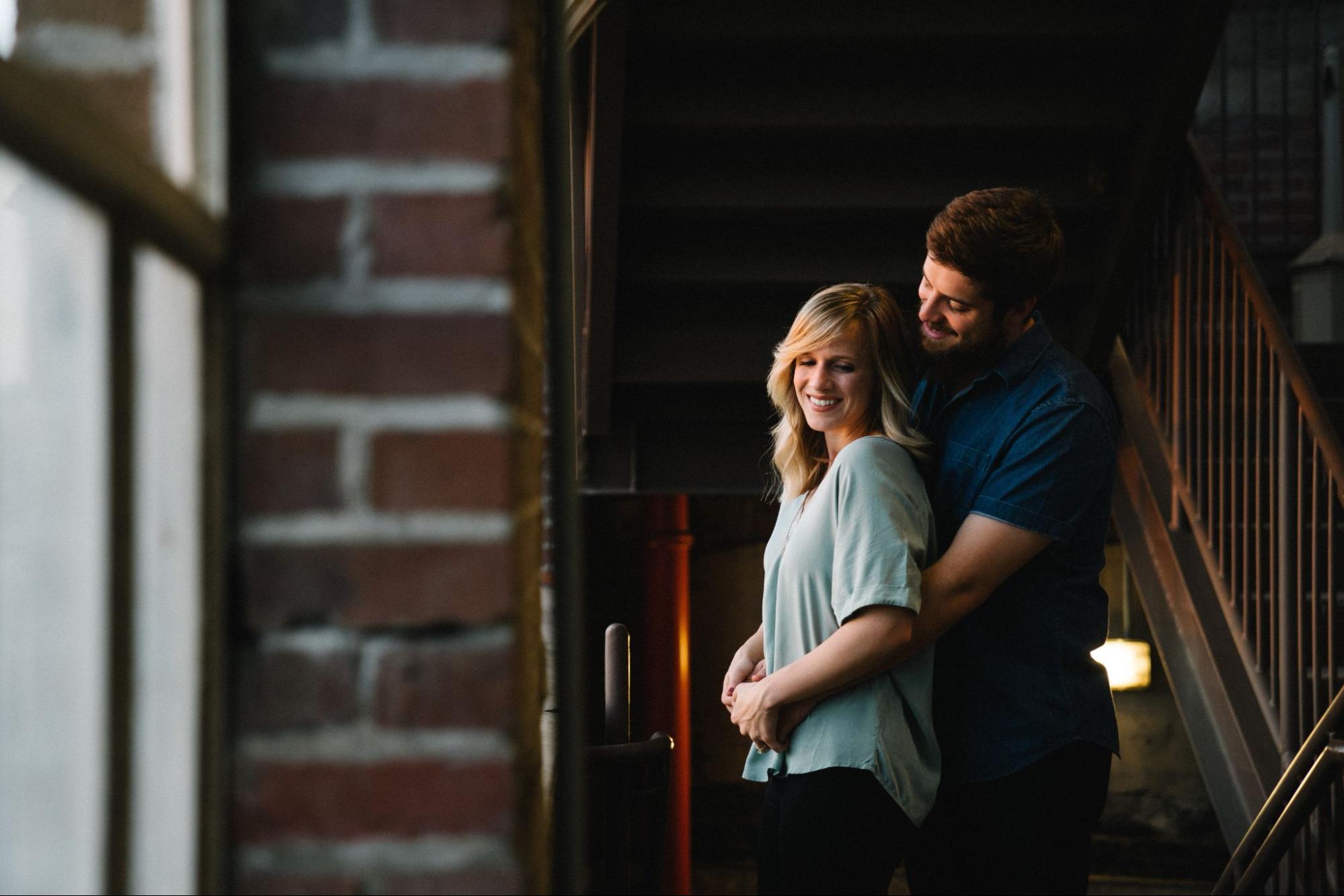 A man standing behind his partner with his hands wrapped around her waist, in a brick room.