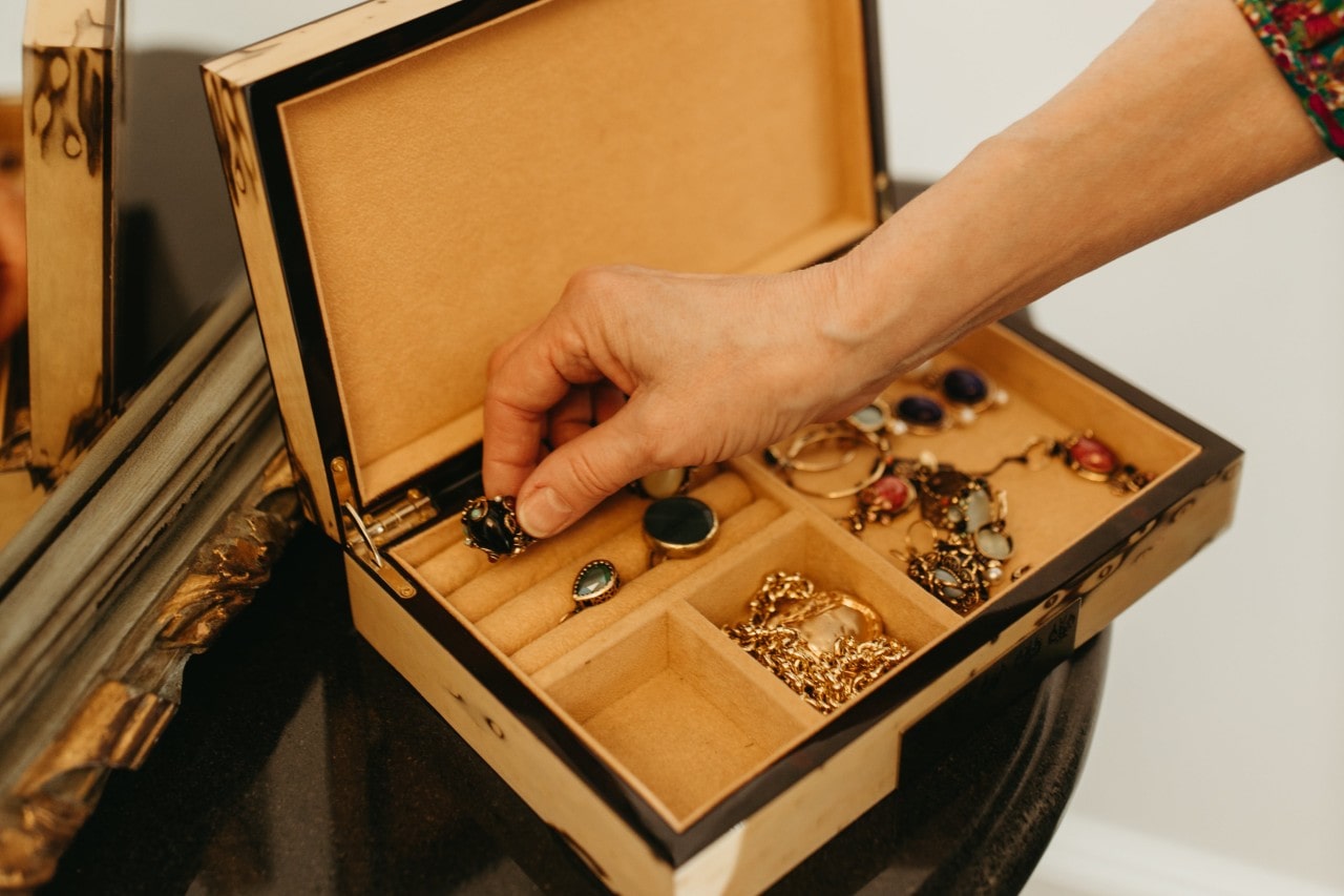 A yellow jewelry box filled with different kinds of jewelry, a hand reaching inside of it to grab a piece.