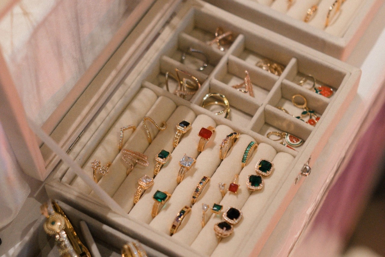 A jewelry box displays arranged gold rings set with colorful gemstones, including emeralds, rubies, and clear stones, organized in soft velvet compartments.