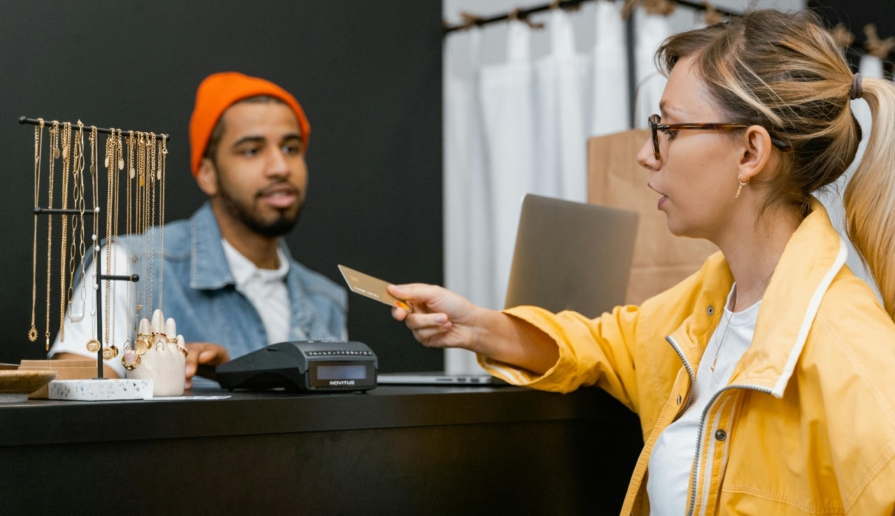 A woman in a yellow jacket holds out a credit card to pay at a jewelry store counter, where a cashier stands behind a display of gold necklaces.