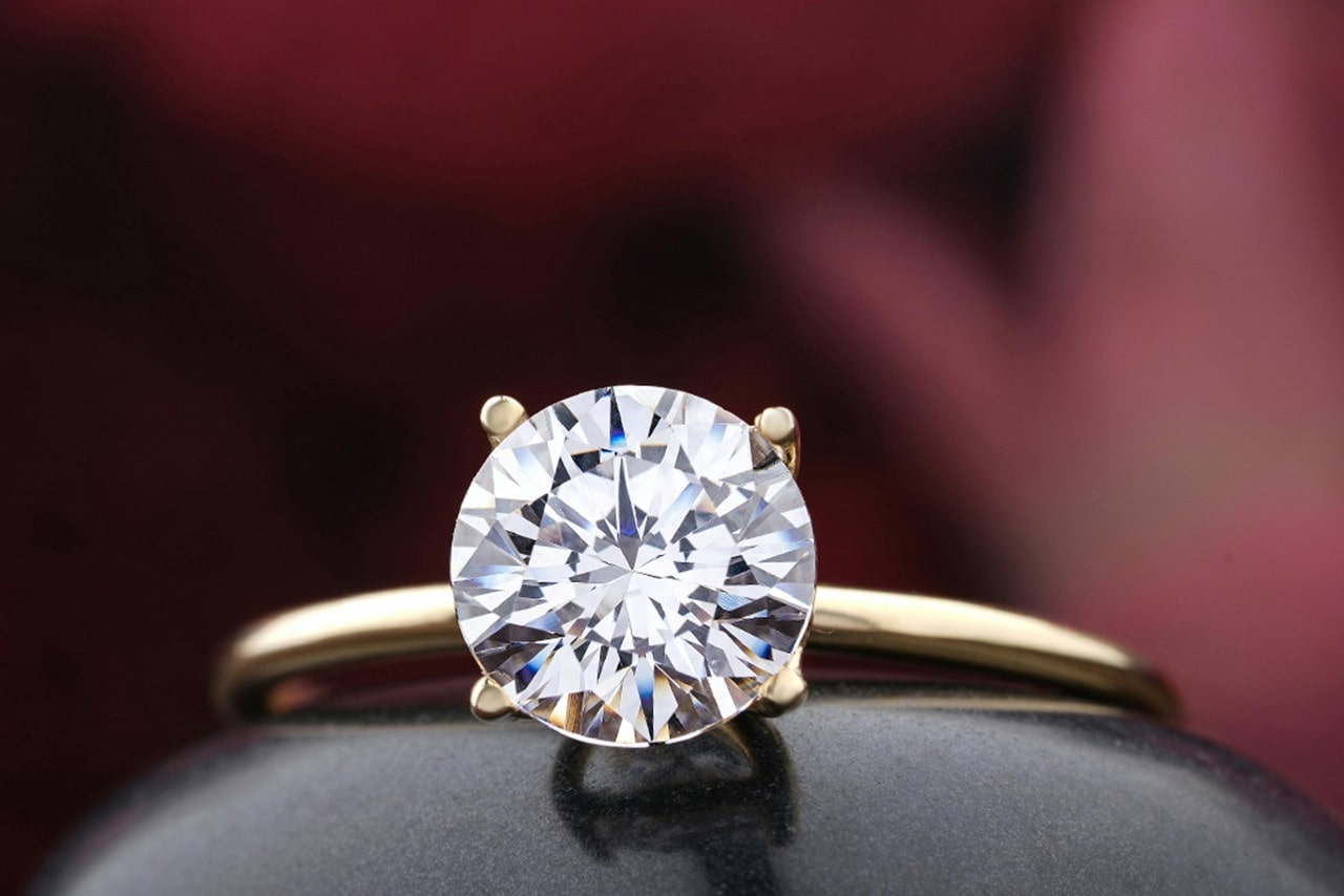 A close-up of a simple yet elegant solitaire engagement ring displayed in front of a crimson backdrop.