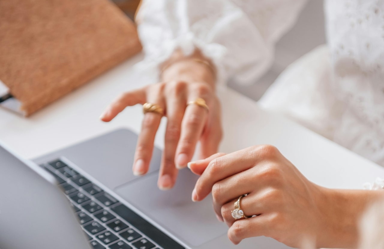 An angled close-up of a woman’s hands adorned with 3 fashion rings as she types on her laptop.