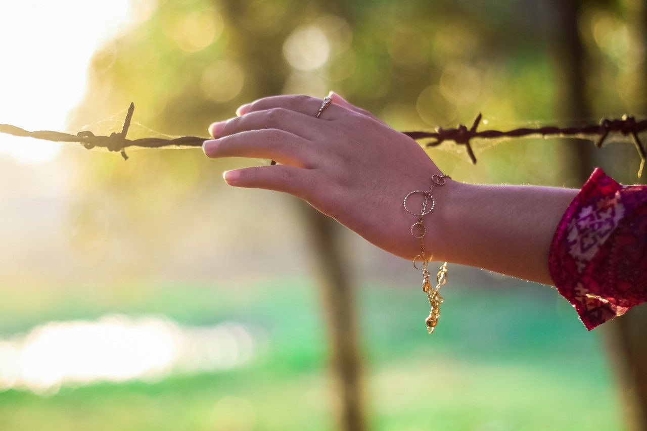 A woman wearing a gold bracelet touches a barbed wire fence at sunset.