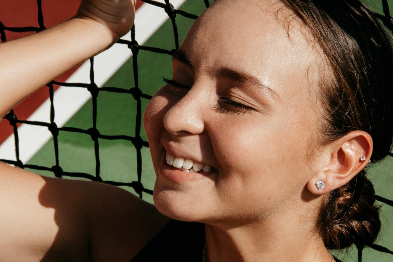 A woman wearing beautiful stud earrings smiles brightly with eyes closed, leaning against a tennis net.