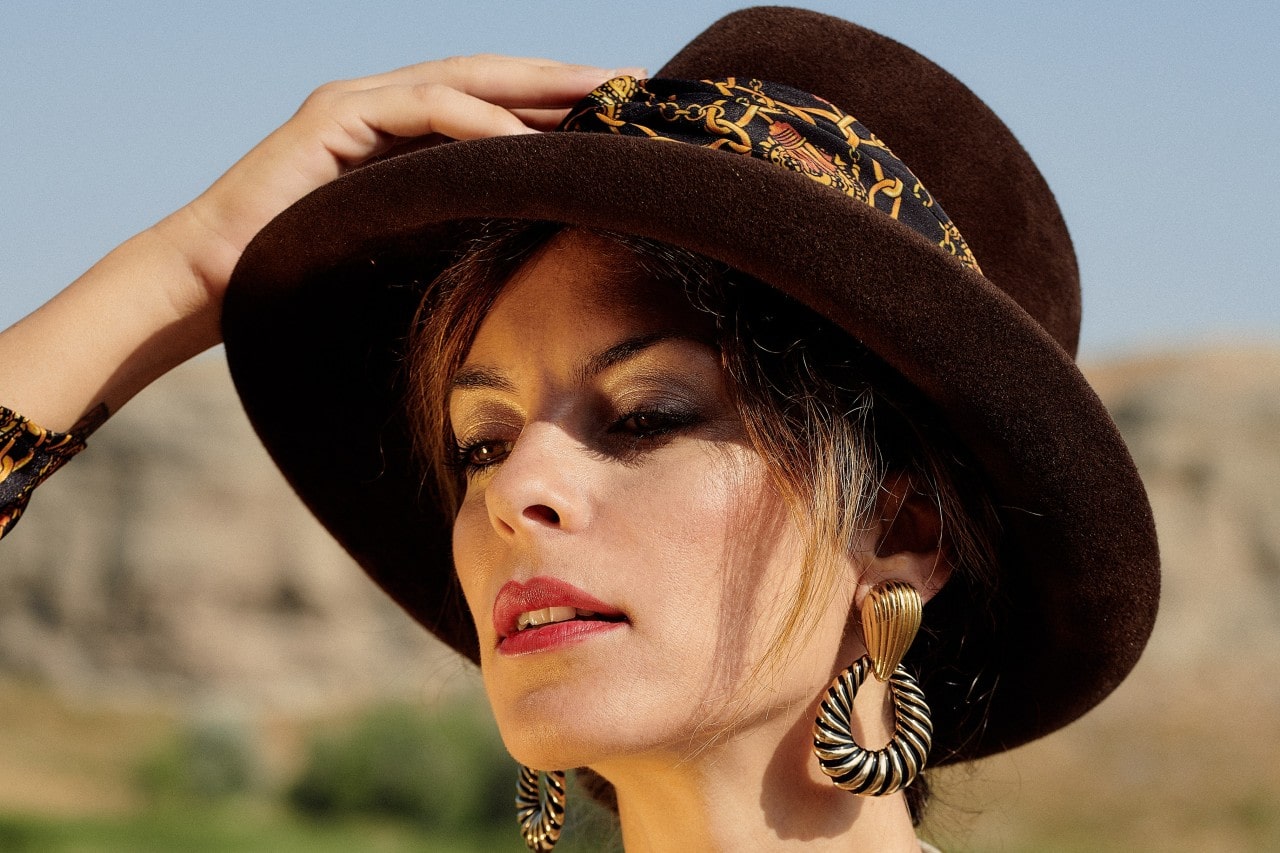 Woman wearing a brown hat with a patterned band, large textured earrings, and subtle makeup.
