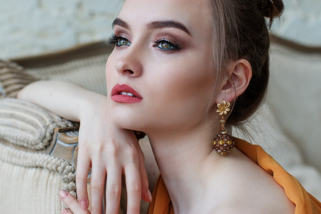A woman with elegant makeup and gold earrings gazes thoughtfully, resting her hand on a beige couch.
