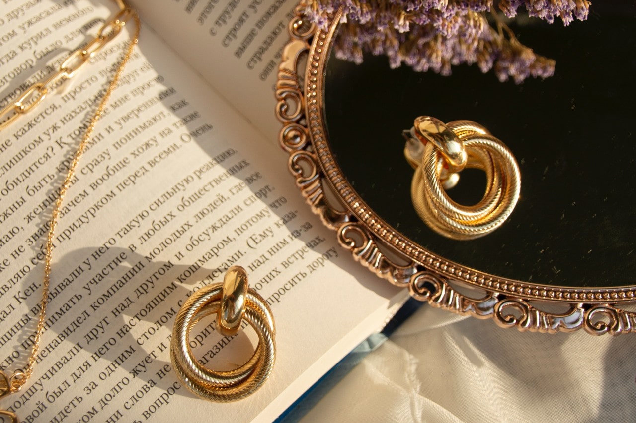 Elegant gold earrings resting on an open book in soft sunlight, reflected in an ornate mirror with lavender flowers.