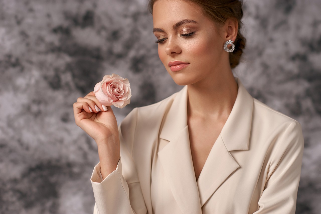 A woman in a beige blazer, holding a rose and wearing white gold diamond round earrings, stands in front of a marble background.