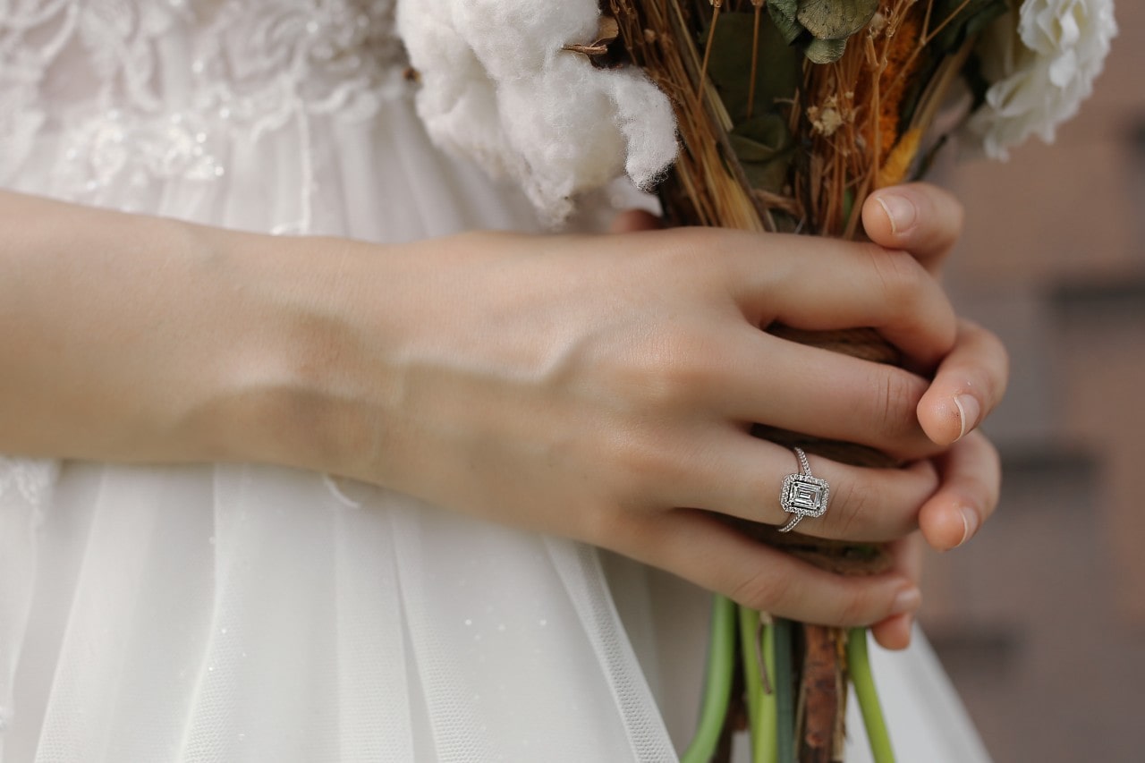 A bride gently holds a bouquet of cotton and dried flowers, showcasing an emerald cut diamond halo engagement ring on her finger. A bride gently holds a bouquet of cotton and dried flowers, showcasing an emerald cut diamond halo engagement ring on her finger.
