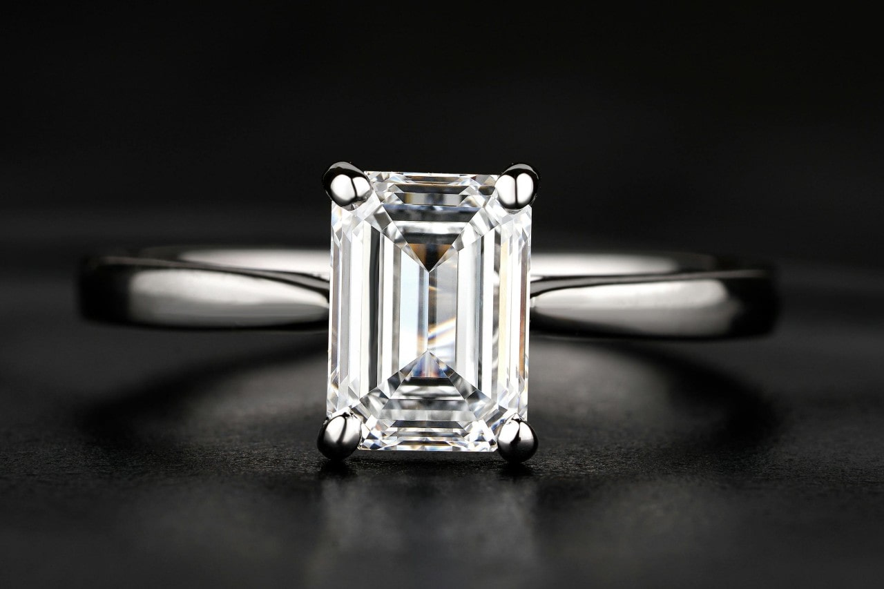A minimalist silver emerald cut solitaire engagement ring on black background. A minimalist silver emerald cut solitaire engagement ring on black background.