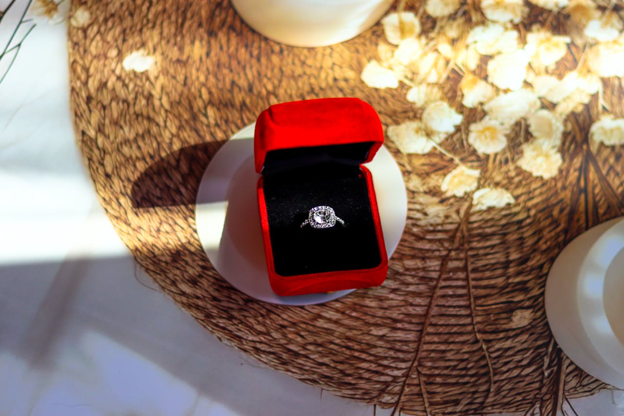 A close up of a white gold halo engagement ring inside a black and red velvet jewelry box on a round woven mat.