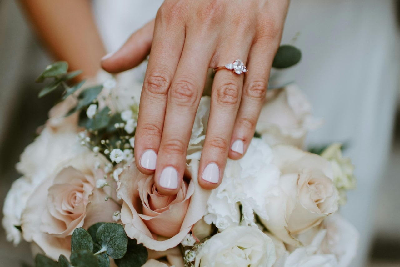 A close-up of a woman’s hand showing an engagement ring while resting on a bouquet of roses and greenery.