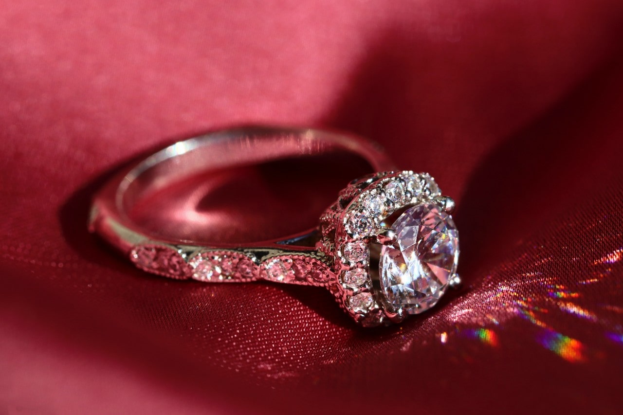 A diamond halo engagement ring with a round brilliant centre stone sparkles on a deep red satin background.