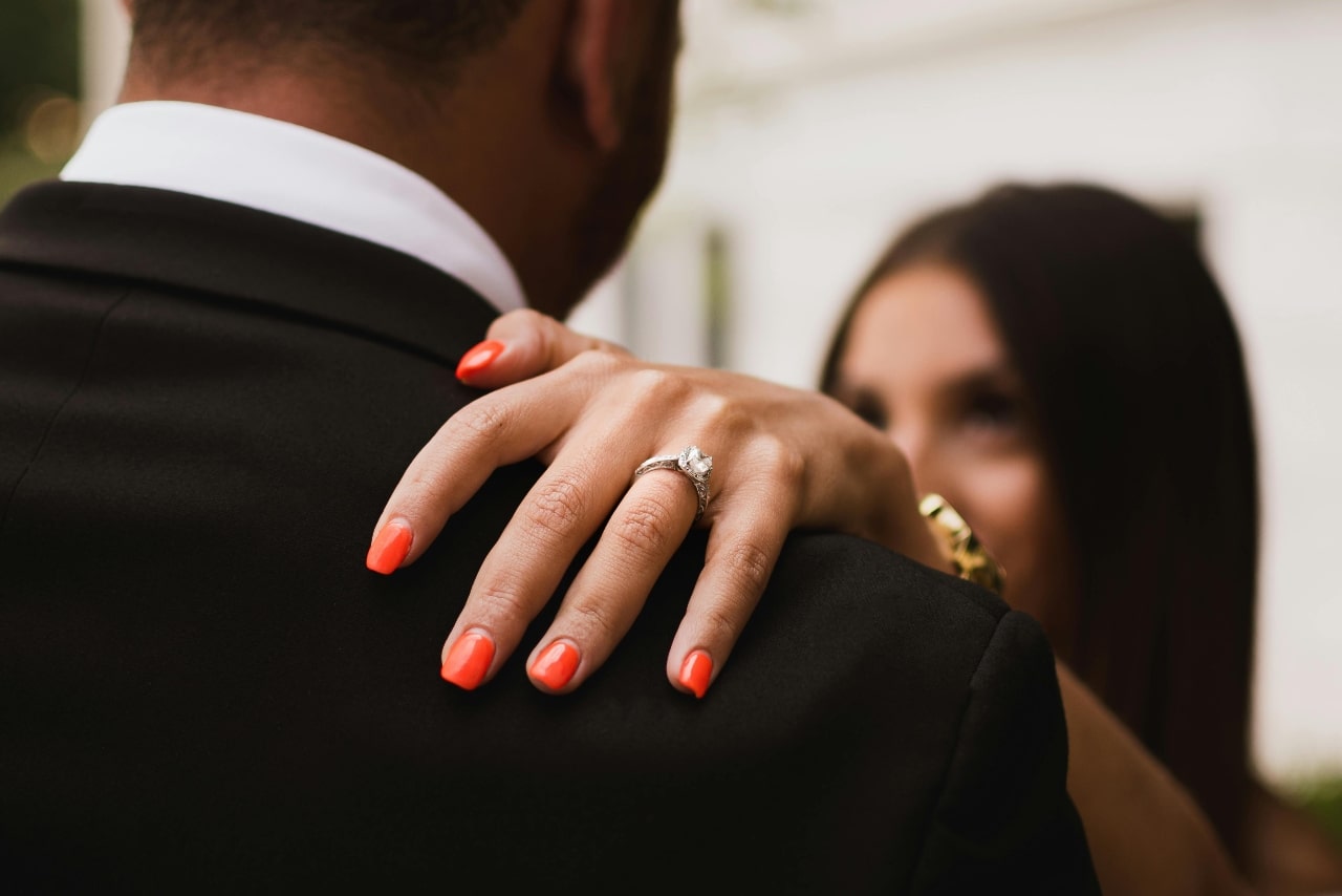 A close up of a woman’s hand with orange manicure and diamond engagement ring on a man’s shoulder in a black suit. A close up of a woman’s hand with orange manicure and diamond engagement ring on a man’s shoulder in a black suit.