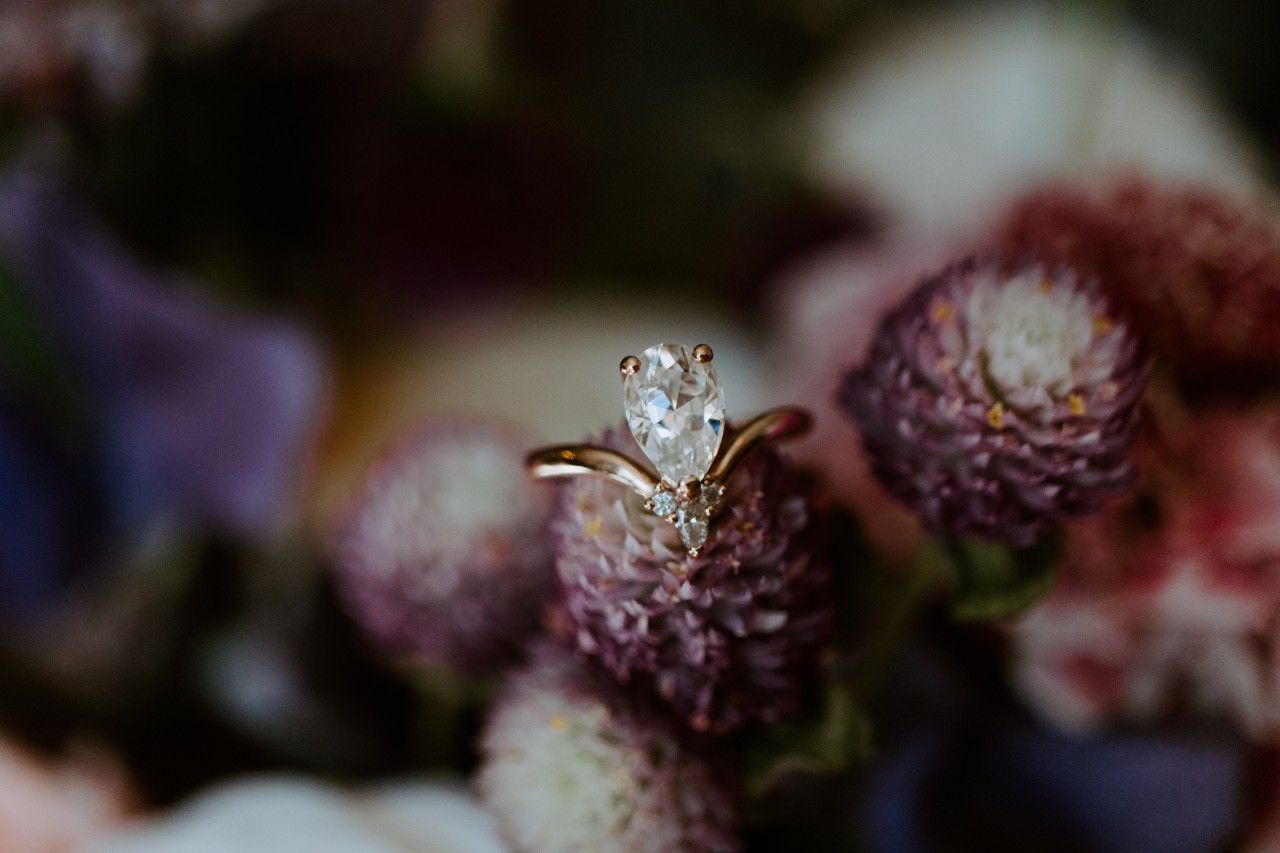 A close up of a v-shaped yellow gold pear-shaped diamond engagement ring against a floral blurry background. A close up of a v-shaped yellow gold pear-shaped diamond engagement ring against a floral blurry background.