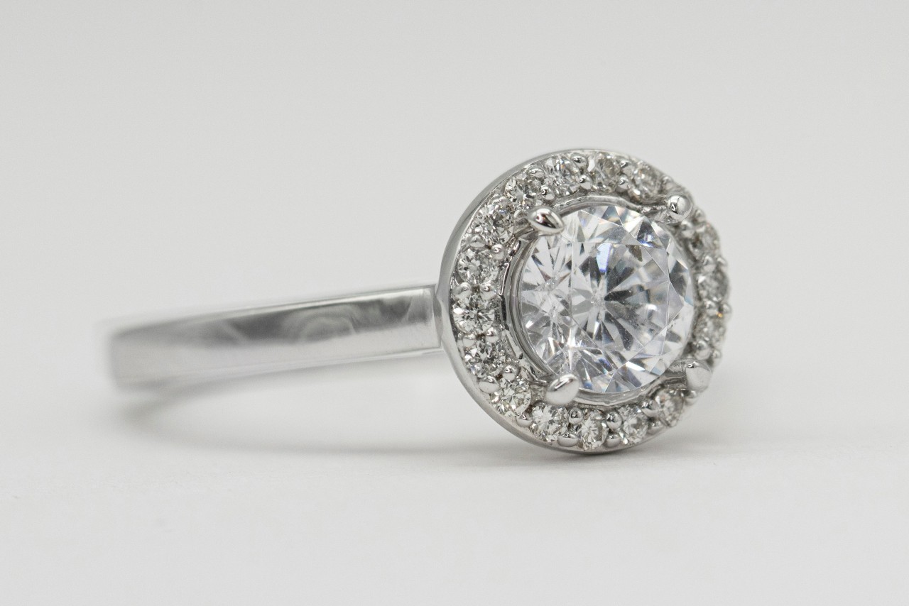 A close-up of a white gold halo engagement ring with a round cut center diamond against a white background. A close-up of a white gold halo engagement ring with a round cut center diamond against a white background.