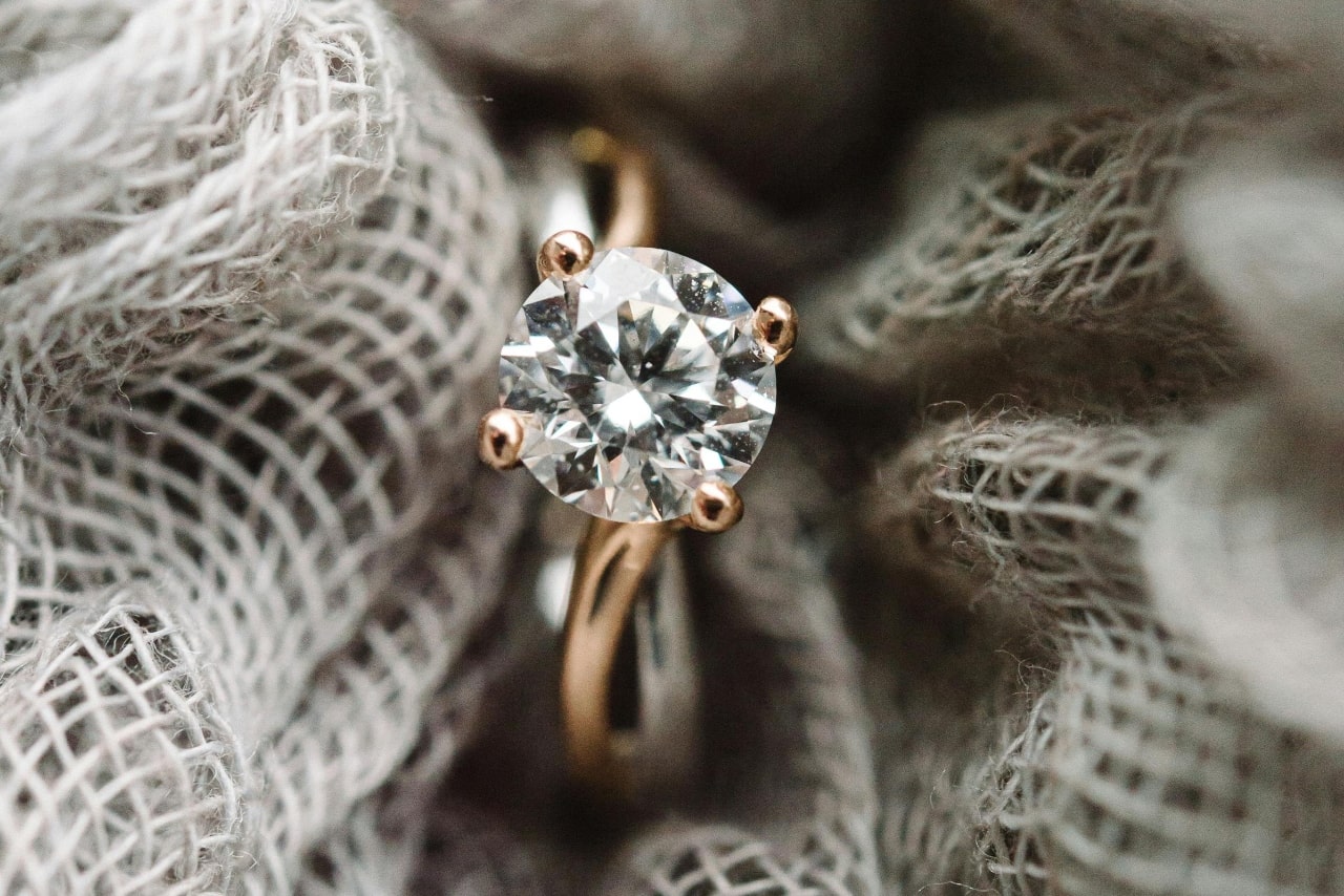 A close-up of a yellow gold solitaire engagement ring with a round center stone held in loosely woven burlap. A close-up of a yellow gold solitaire engagement ring with a round center stone held in loosely woven burlap.