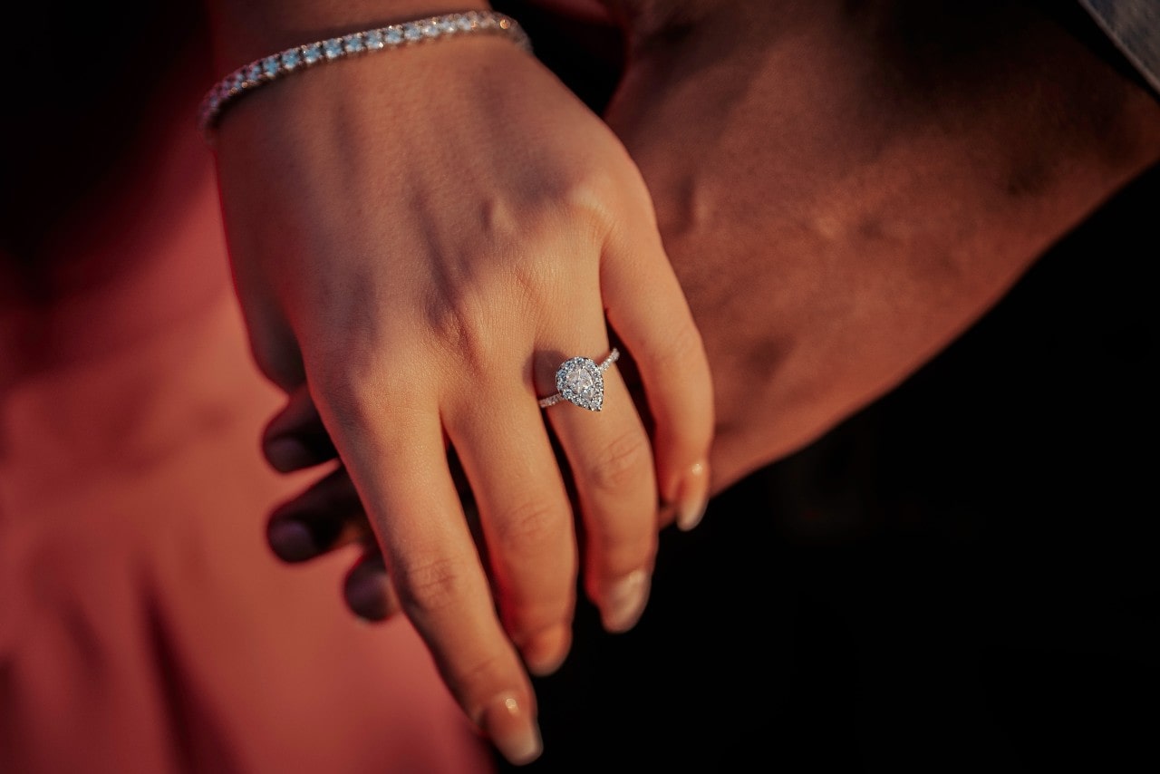 A close-up of a bride’s hand atop the groom’s, a white gold halo engagement ring with a pear cut center diamond on her finger. A close-up of a bride’s hand atop the groom’s, a white gold halo engagement ring with a pear cut center diamond on her finger.