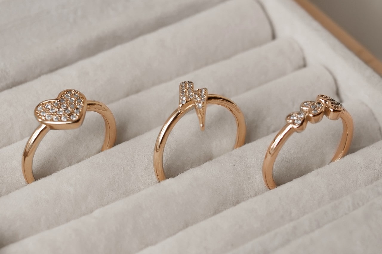 A close up of 3 yellow gold fashion rings with sparkling diamonds in a different shape – a heart, a lightning bolt, and a trio of round stones – rest in individual slots within a light-colored jewelry display. A close up of 3 yellow gold fashion rings with sparkling diamonds in a different shape – a heart, a lightning bolt, and a trio of round stones – rest in individual slots within a light-colored jewelry display.