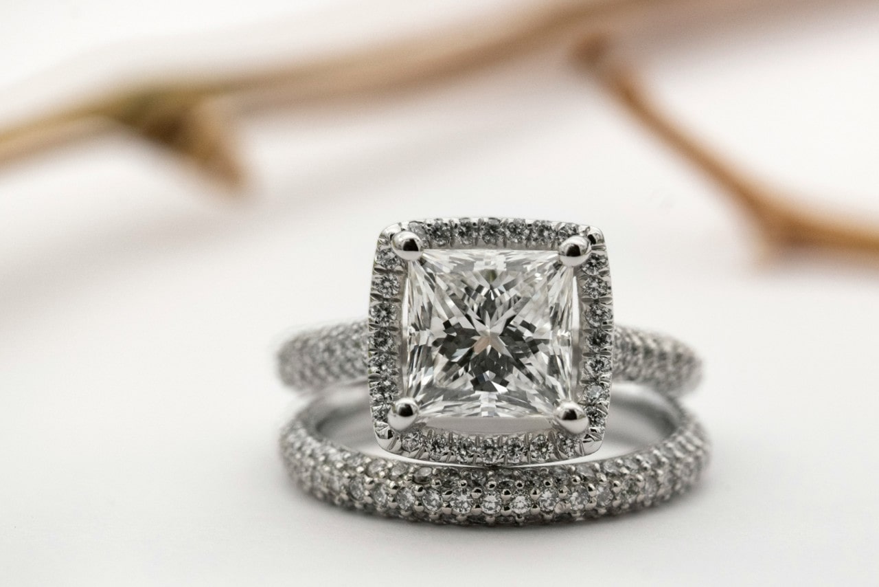 A close-up of an ornate bridal set, the engagement ring featuring a halo design with a cushion cut center stone.