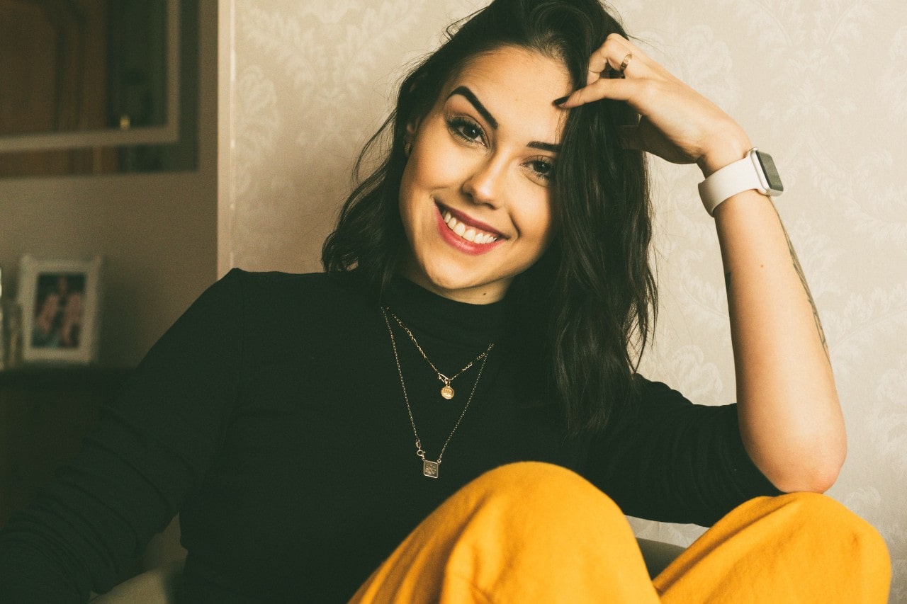 A smiling woman in a black top and yellow pants, seated casually, wearing layered necklaces and a watch.
