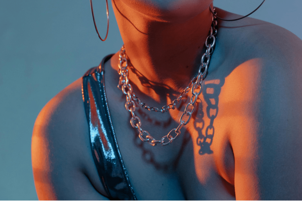 A close-up of a person wearing large hoop earrings, layered chain necklaces, and a shiny blue top.