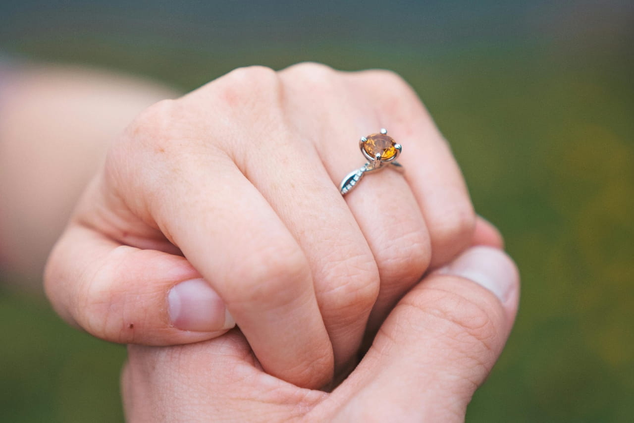 A woman resting her hand in her partner's, showcasing her amber colored engagement ring with a silver band.