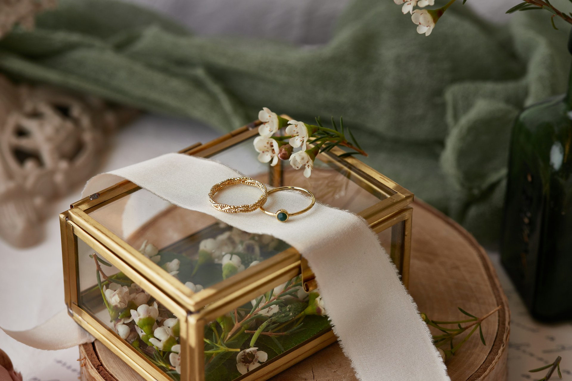 Two gold wedding bands rest on a white ribbon atop a golden glass box adorned with small white flowers and a green ribbon.