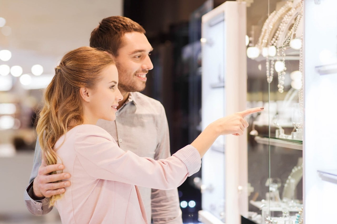 Couple shopping for jewelry; the woman gestures toward a piece while the man stands behind her with a hand on her back.