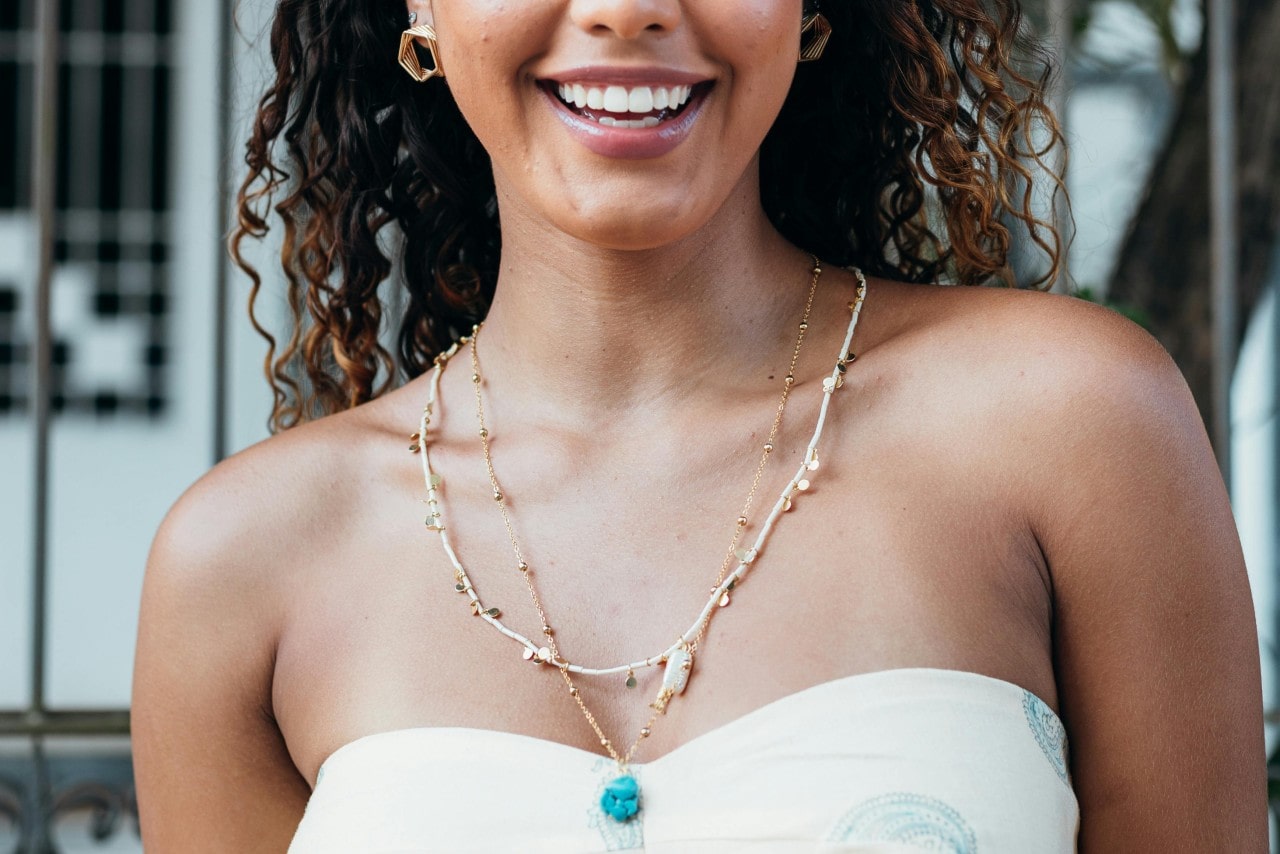 Woman with curly hair wearing a layered yellow gold necklace featuring a blue gemstone at the center.