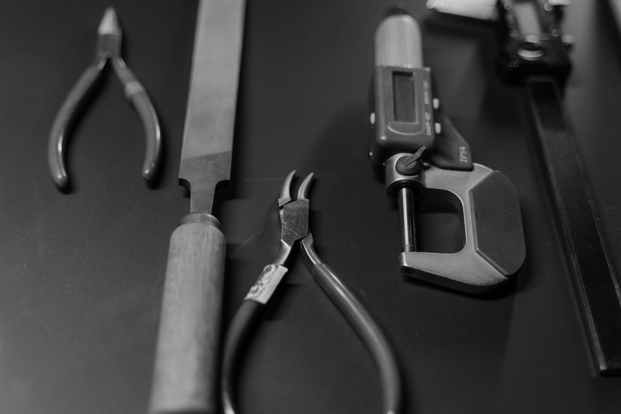 A black and white close-up of jeweler’s tools, including pliers and a file, atop a work desk. A black and white close-up of jeweler’s tools, including pliers and a file, atop a work desk.