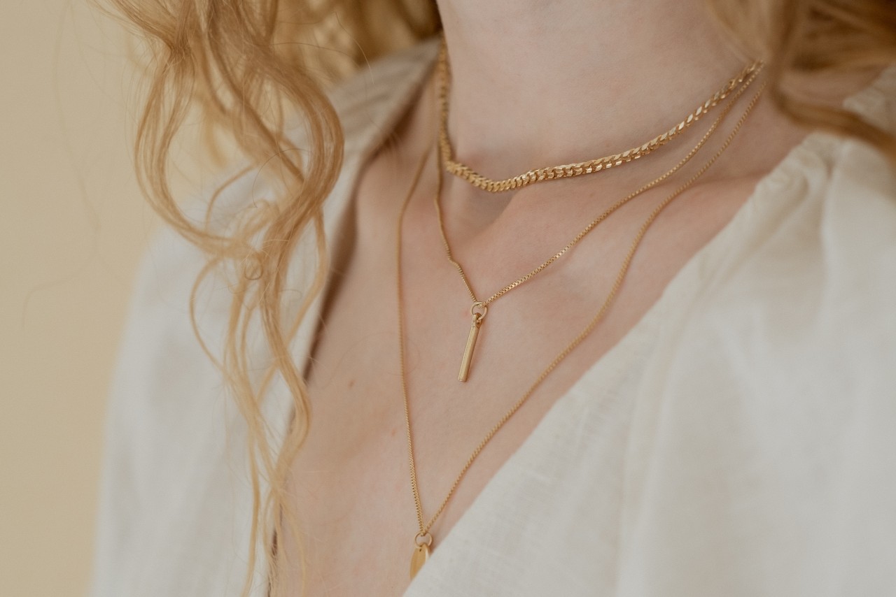 A close-up of a woman’s shoulders and chest in a plunging white blouse, her collarbone adorned with three layered gold chains. A close-up of a woman’s shoulders and chest in a plunging white blouse, her collarbone adorned with three layered gold chains.